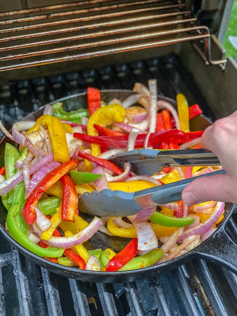 peppers and onions being tossed with tongs in cast iron skillet on the grill
