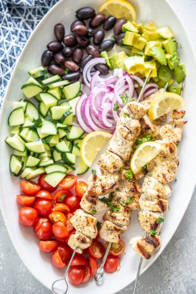 oval platter filled with chicken kabobs, tomatoes, cucumbers, olives and onions